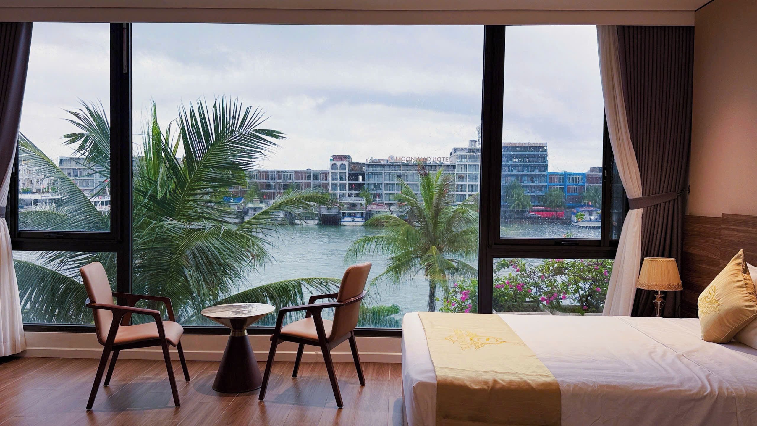 Habitación de hotel con un ventanal grande, una cama, dos sillas, una mesita y vistas a un río y edificios.