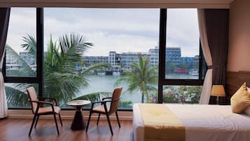 Habitación de hotel con un ventanal grande, una cama, dos sillas, una mesita y vistas a un río y edificios.