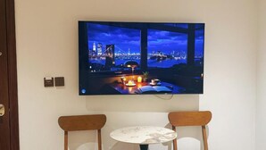 Et fladskærms-tv på væggen viser en by nattebelysning med en bro. Under tv'et står et rundt bord med to stole.