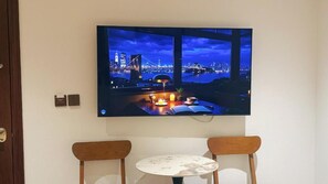 Un televisor de pantalla plana montado en la pared muestra un paisaje urbano nocturno con un puente. Debajo del televisor, hay una mesa redonda pequeña con dos sillas.