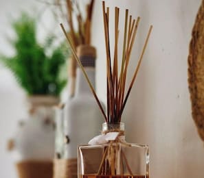 A clear glass bottle with reed sticks and a white cloth, possibly a diffuser, on a surface with a blurred background.