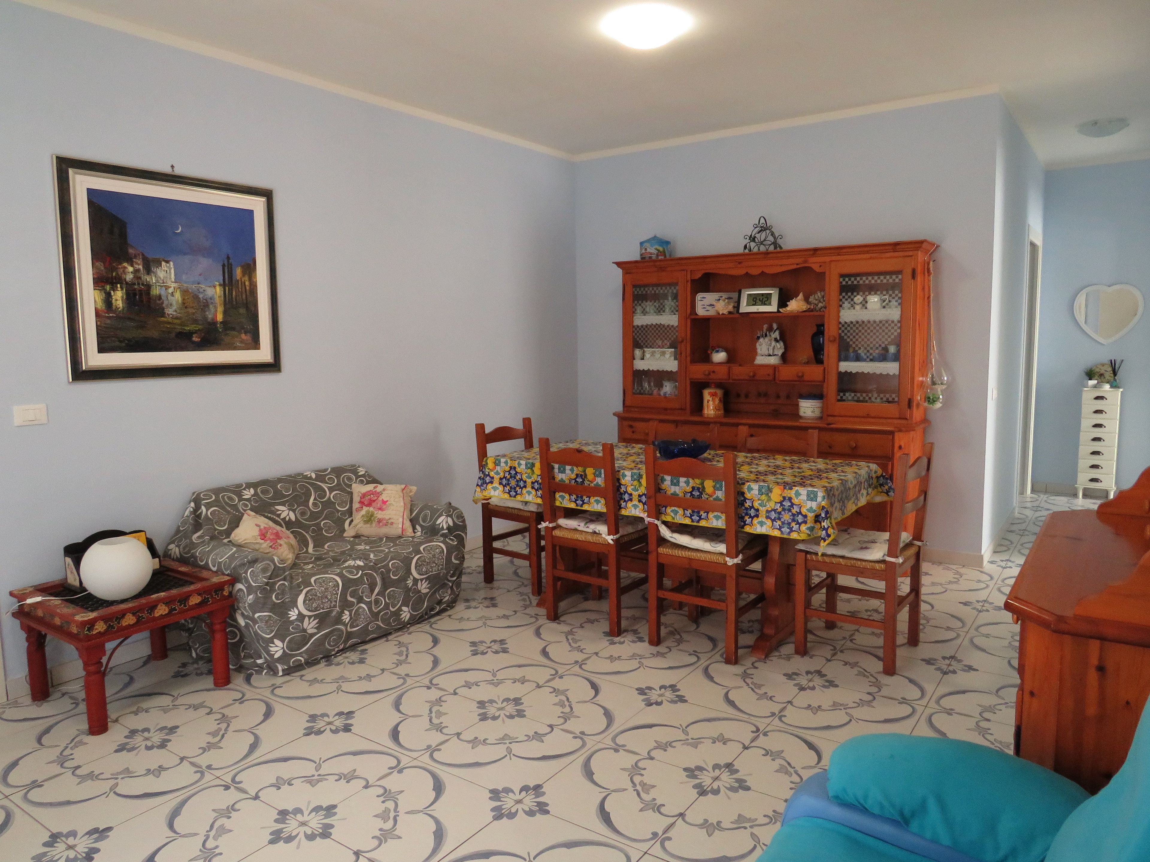 Una stanza con un pavimento a motivi, un divano, un tavolo da pranzo con sedie, un mobile contenitore con oggetti decorativi e un quadro appeso al muro.
