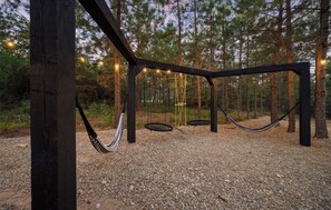 A hammock suspended between wooden beams in a forested area with string lights. - Modern Hochatown Cabin - Hot Tub, 2 Kings, Arcade + Firepit (Broken Bow)