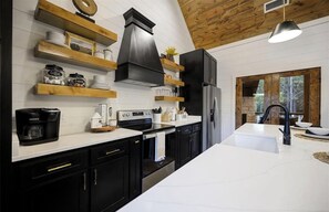 A modern kitchen with black cabinets, white countertops, and wooden shelves. - Modern Hochatown Cabin - Hot Tub, 2 Kings, Arcade + Firepit (Broken Bow)