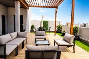 Un área de estar moderna al aire libre con sofás, sillones y una mesa, rodeada de plantas en macetas y una pérgola de madera.