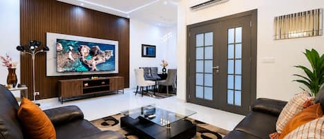 A modern living room with a flat-screen TV, leather sofas, a glass coffee table, and a dining area with a chandelier.