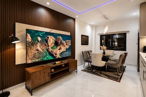 A modern living room with a large flat-screen TV, wooden paneling, a dining area with a glass table, and a kitchen with a black countertop. - Spacious 3-bedroom apartment Dakar Fast Wifi with AC and gym, Centrally Located (Dakar)