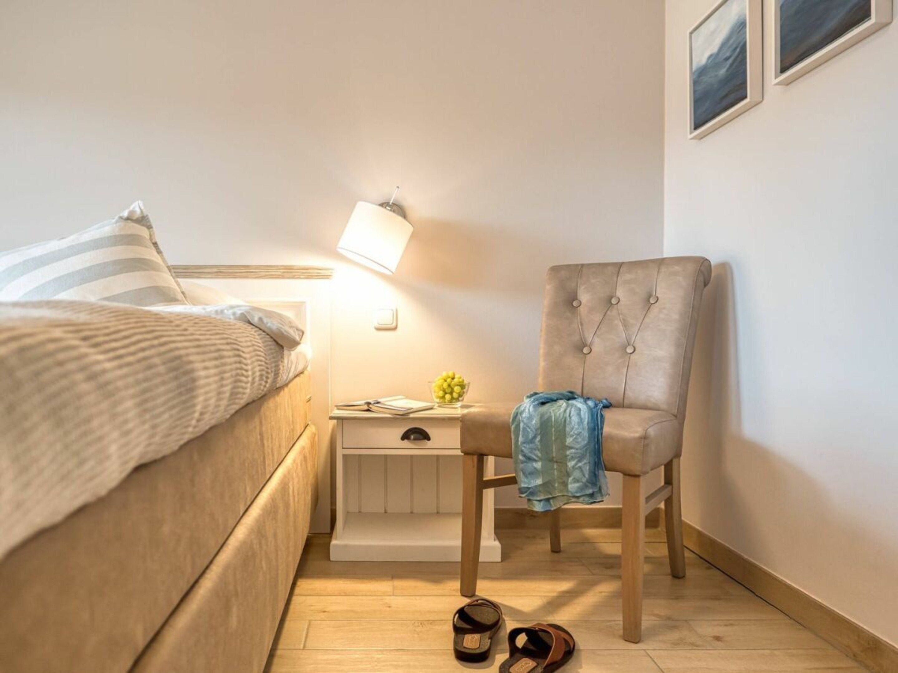 A bedroom with a bed, bedside table, lamp, chair, and a pair of sandals on the floor.