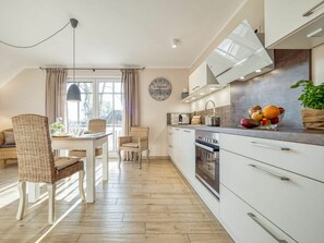 Eine moderne Küche mit weißen Schränken, eine Essecke mit Korbstühlen und ein großes Fenster mit Vorhängen.