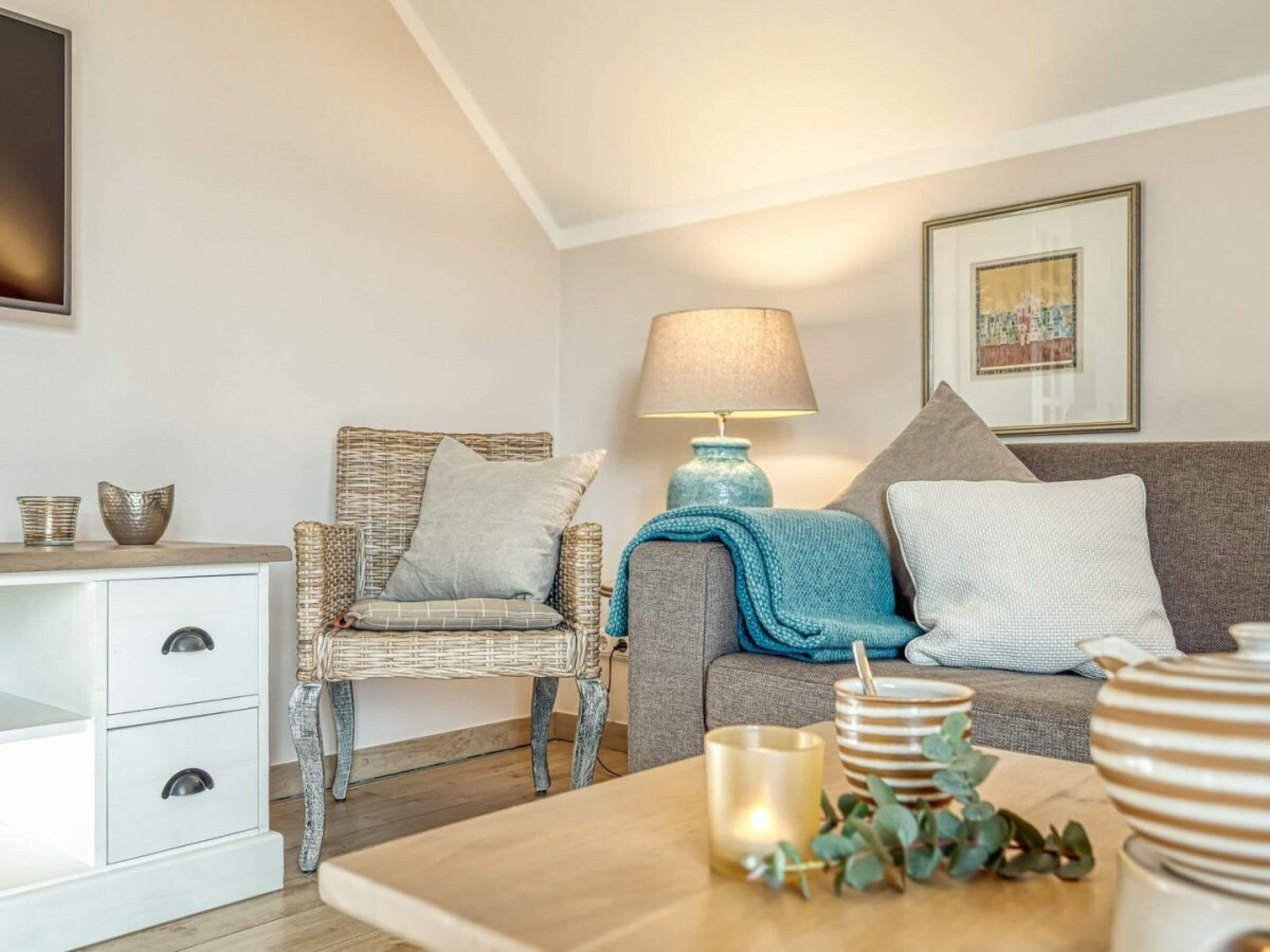 Een gezellige woonkamer met een rieten stoel, een grijze bank, een wit kastje, een blauwe lamp en een ingelijst kunstwerk aan de muur.