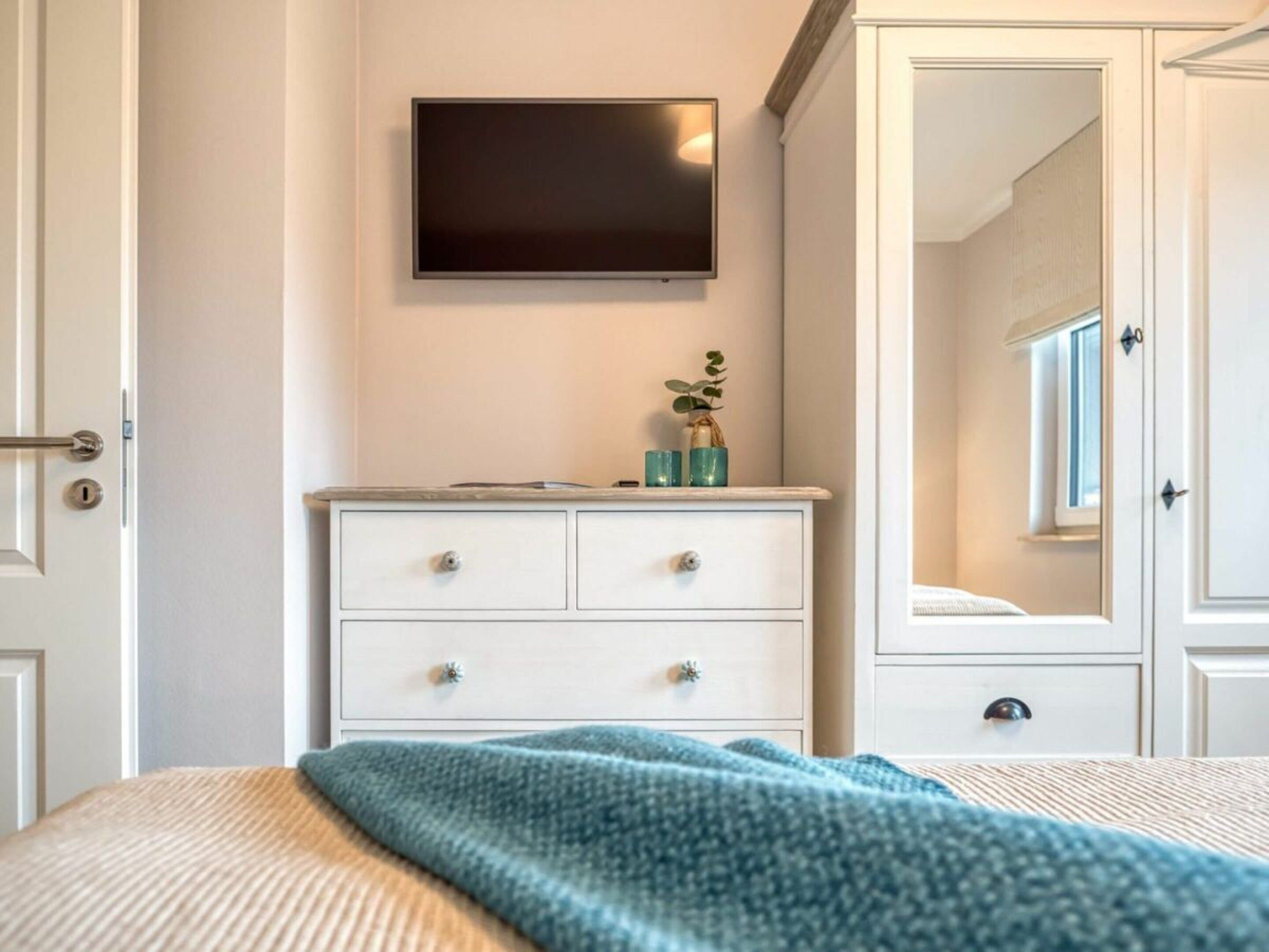 Quarto com cama, cômoda, televisão na parede e espelho.