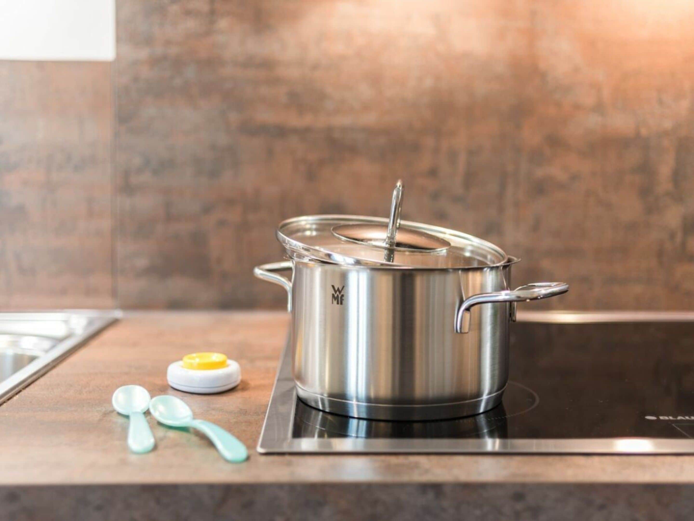 A stainless steel pot on an induction cooktop with a lid, a yellow and white container, and two blue spoons on a kitchen counter.