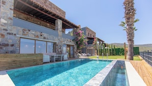 A swimming pool with a stone wall and a wooden deck, a lounge area with chairs, and a view of the surrounding landscape. - mira villa 2 (Torba)