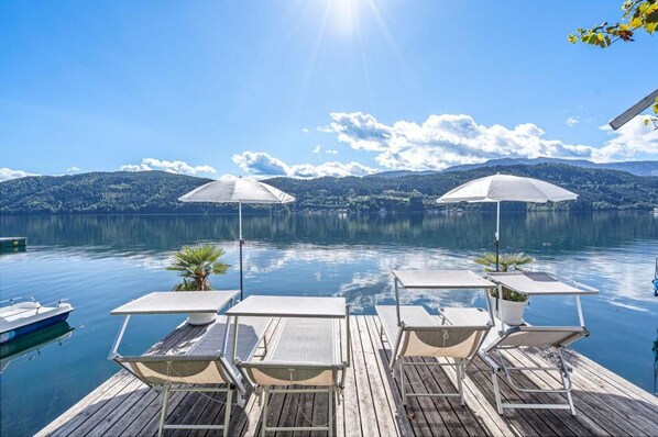Terraza de madera con sillones de descanso y sombrillas frente a un lago tranquilo con montañas al fondo.