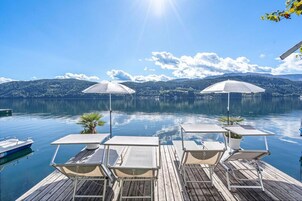 Um deck de madeira com espreguiçadeiras e guarda-sóis, com vista para um lago tranquilo e montanhas ao fundo.