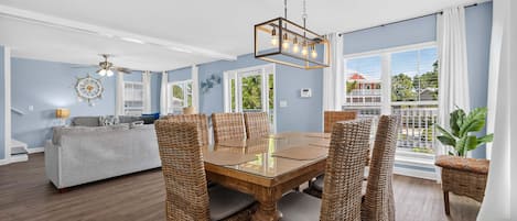 A dining area with a wooden table and wicker chairs, a ceiling fan, and a view of the outdoors through large windows.