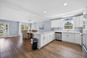 A modern kitchen with white cabinetry, a dining area with wicker chairs, and a view of the outdoors through a large window. - The Great Escape (Gulf Shores)