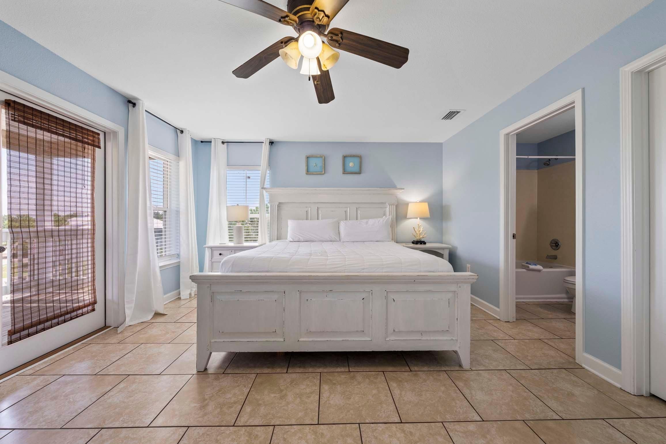 A bedroom with a large bed, a ceiling fan, a window with blinds, and a bathroom visible through an open door.