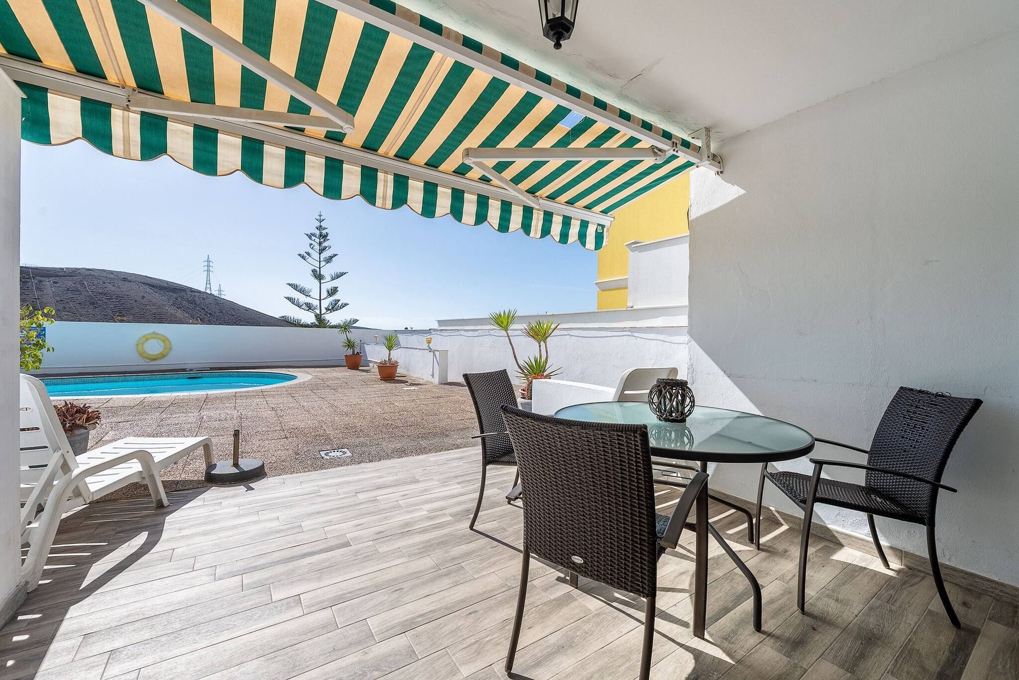 En udendørs terrasse med swimmingpool, et bord med stole og en stribet markise.