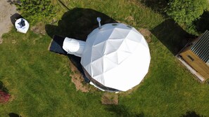 Vista aérea de uma grande estrutura branca em formato de tenda em um campo gramado, com um pequeno edifício ao fundo.