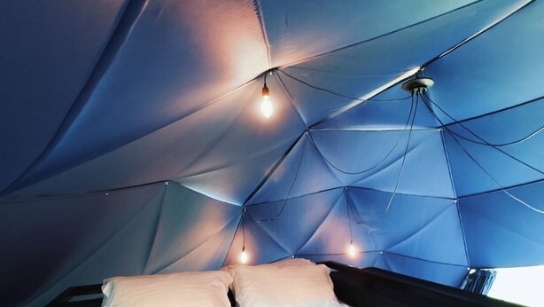 A tent-like interior with a bed, multiple light bulbs, and a window. - Luxury Igloo Dome with Hot Tub (Redberth)