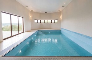 Ein großer, überdachter Schwimmbereich mit klarem, blauem Wasser, gefliestem Boden und Blick ins Freie durch große Fenster.