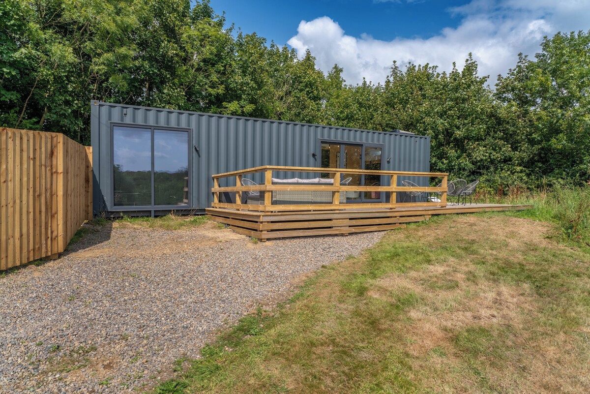 A compact cabin with a deck, a large sliding door, and a gravel driveway.