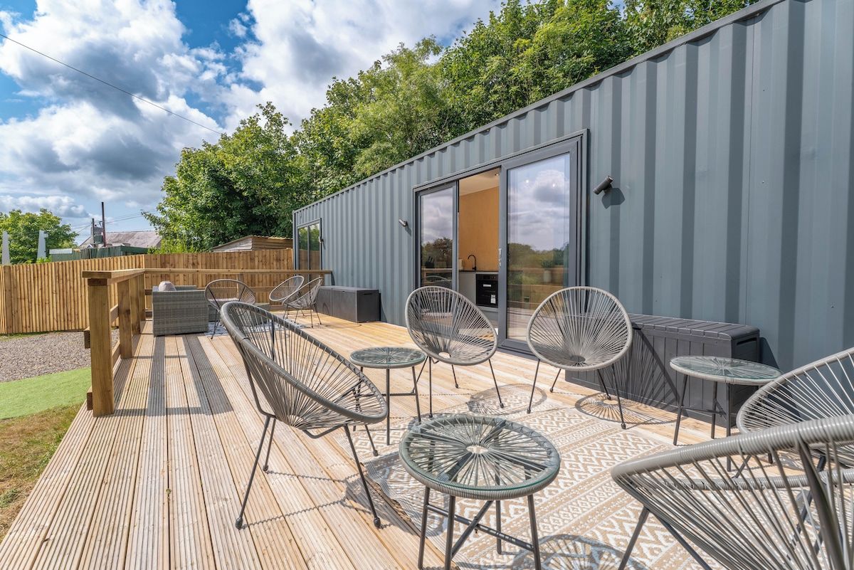 An outdoor deck with wicker chairs and tables, a wooden fence, and a grey building with a sliding glass door.