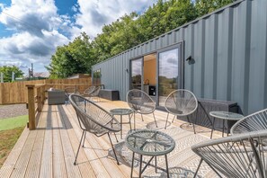 An outdoor deck with wicker chairs and tables, a wooden fence, and a grey building with a sliding glass door. - Modern Tiny Home with Hot Tub (Redberth)