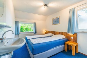 A hotel room with a bed, a bedside table, a window with curtains, a wall-mounted light, and a bathroom with a sink and mirror. - Charming Villa With Enclosed Garden (De Koog)