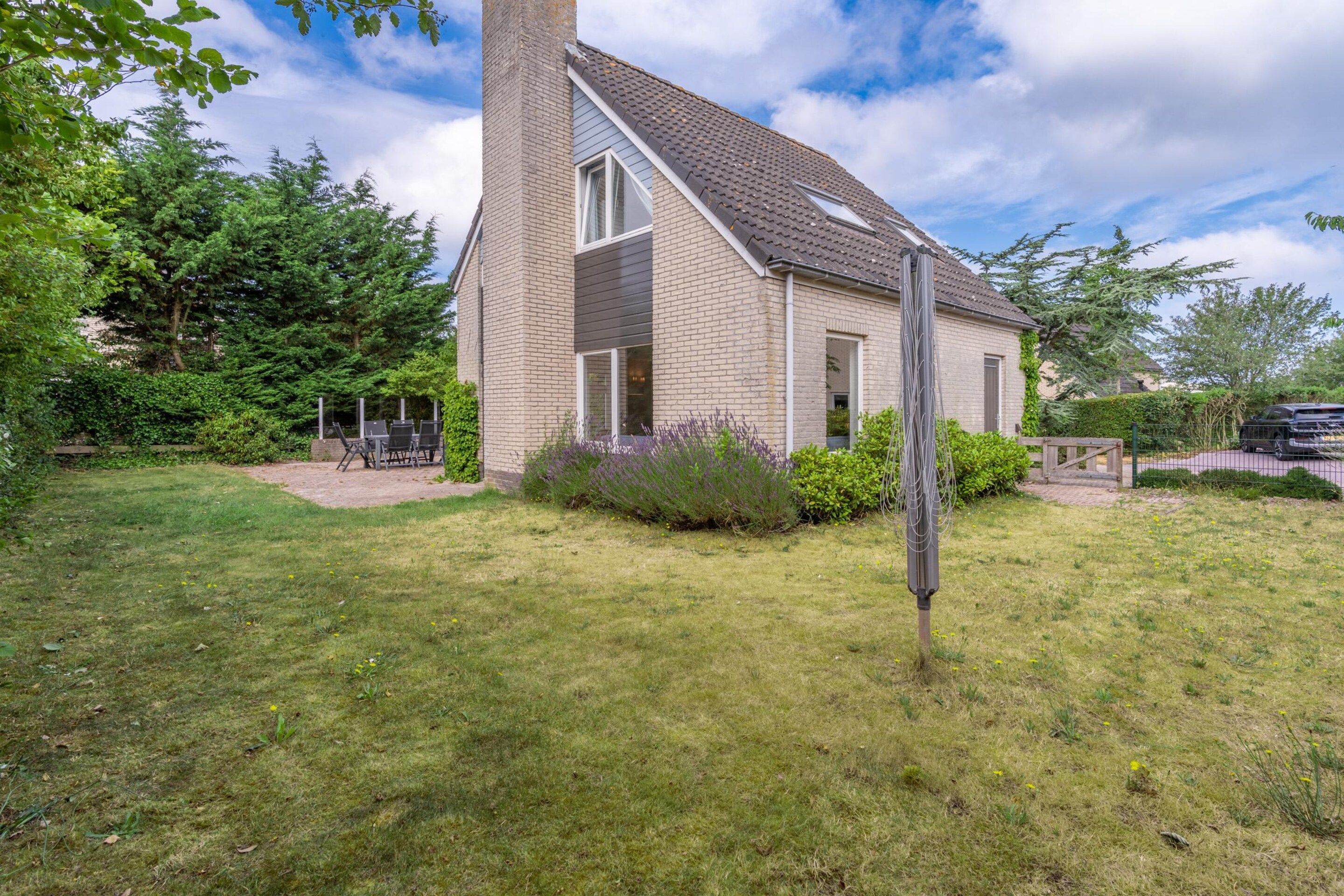 A two-story house with a visible chimney, a garden with grass and shrubs, and a car parked in the driveway.