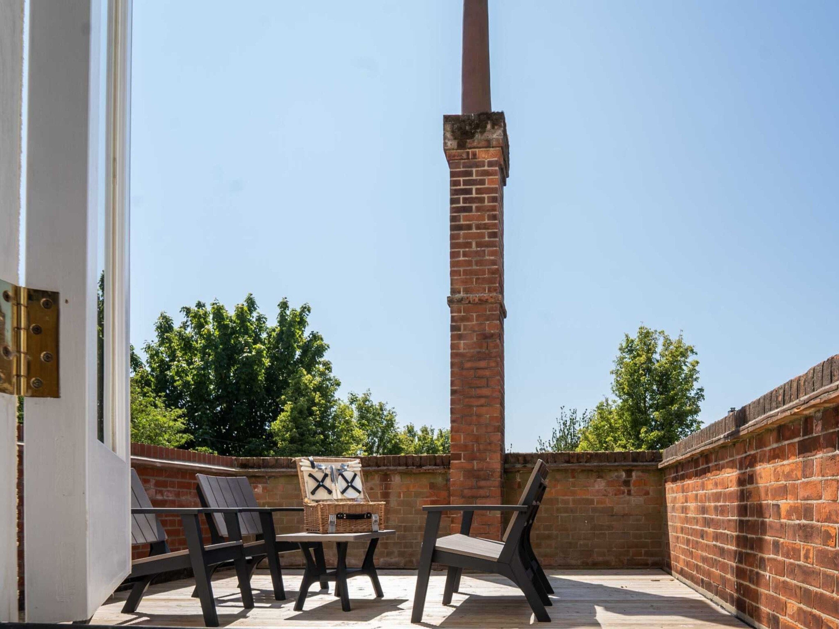 An outdoor patio with a brick chimney, two wooden chairs, a small table, and a wicker basket.