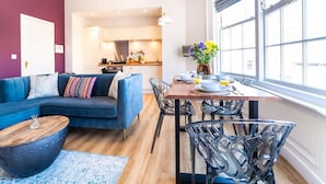 Een moderne woonkamer met een blauwe bank, een eettafel met stoelen, en een keuken op de achtergrond.