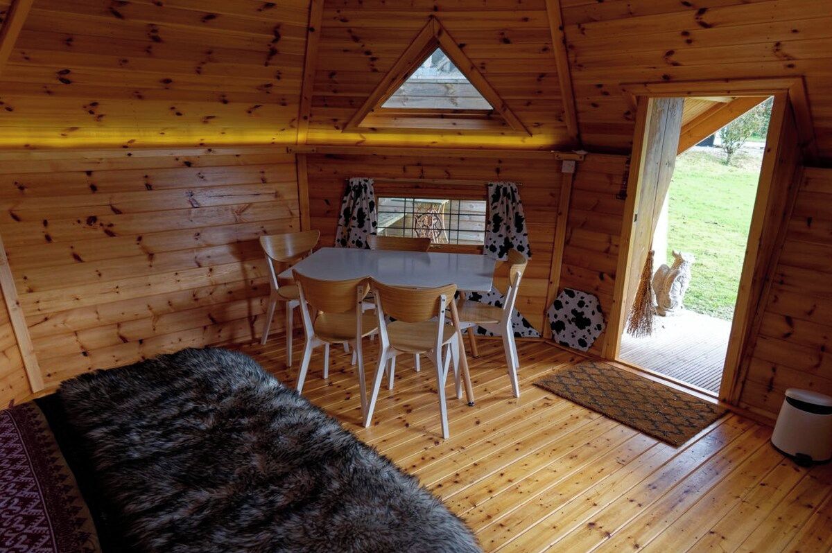 A cozy cabin interior with a wooden dining table and chairs, a fur-covered bench, and a door leading outside.