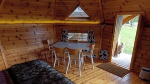 Interior acogedor de una cabaña con una mesa y sillas de madera, un banco cubierto de pieles y una puerta que da al exterior.