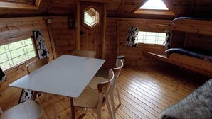 Interior de una cabaña con una mesa de comedor, sillas, literas y ventanas con cortinas.