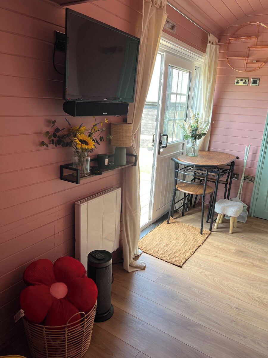 A cozy room with a television mounted on the wall, a dining table with chairs, a radiator, and a basket with red cushions.