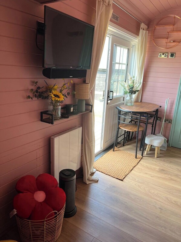 A cozy room with a television mounted on the wall, a dining table with chairs, a radiator, and a basket with red cushions.