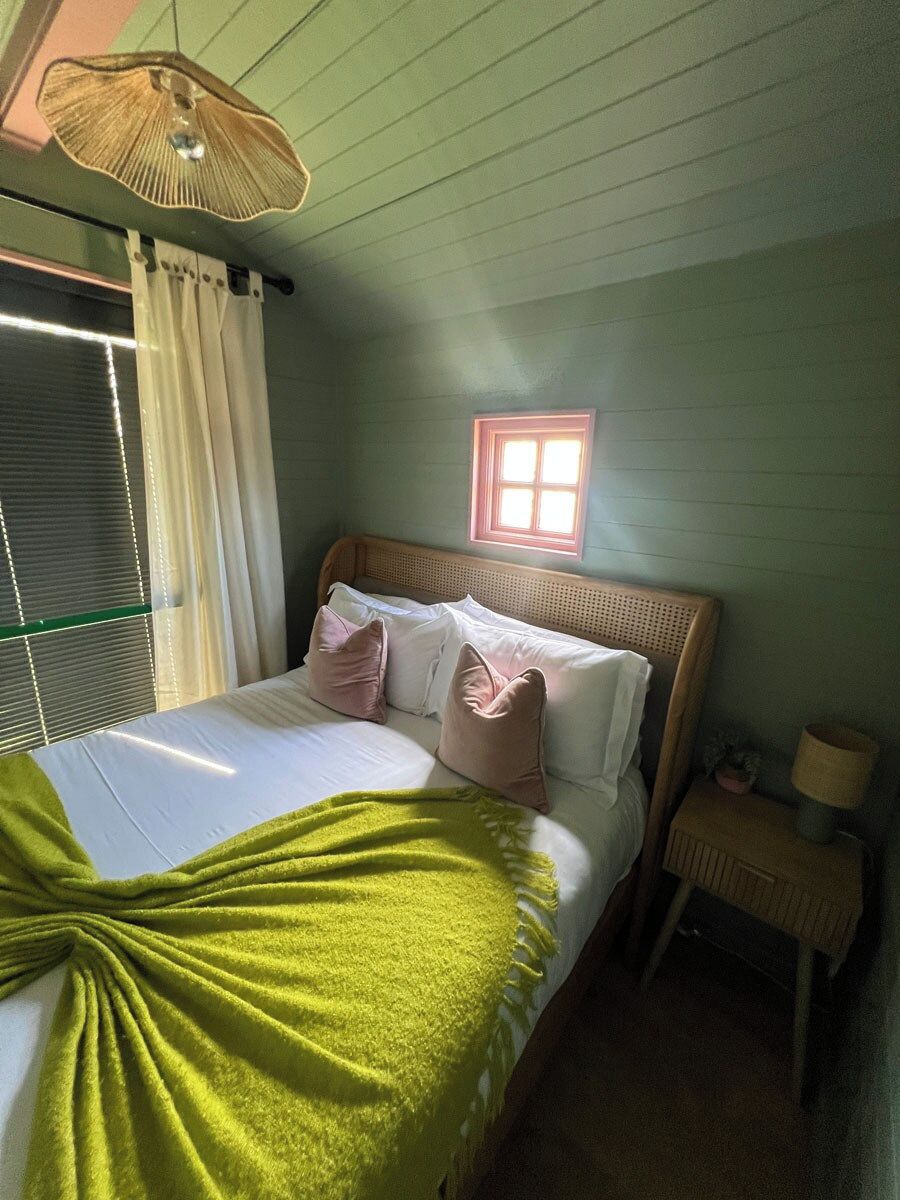 Un dormitorio con cama, almohadas, una mesita de noche, lámpara, ventana y ventilador de techo.