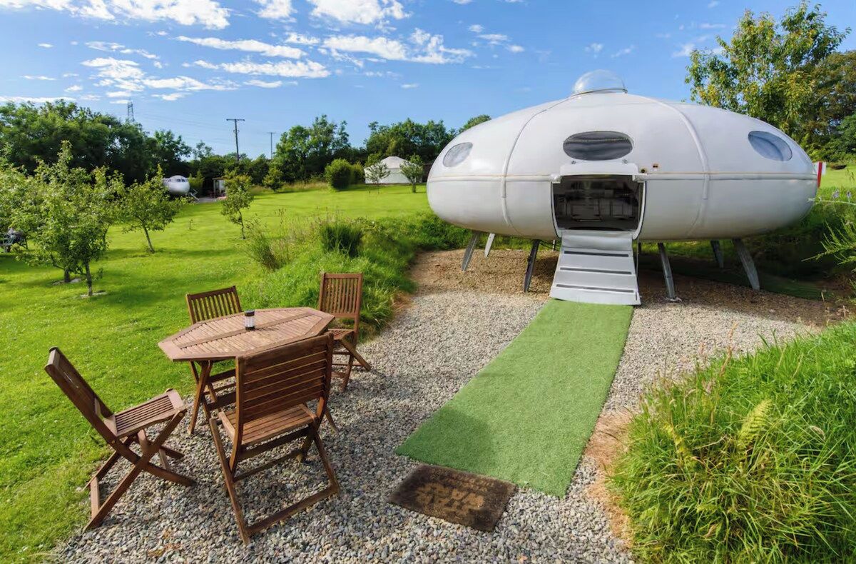 A geodesic dome structure with a small ramp leading to an entrance, surrounded by a grassy area with a table and chairs.