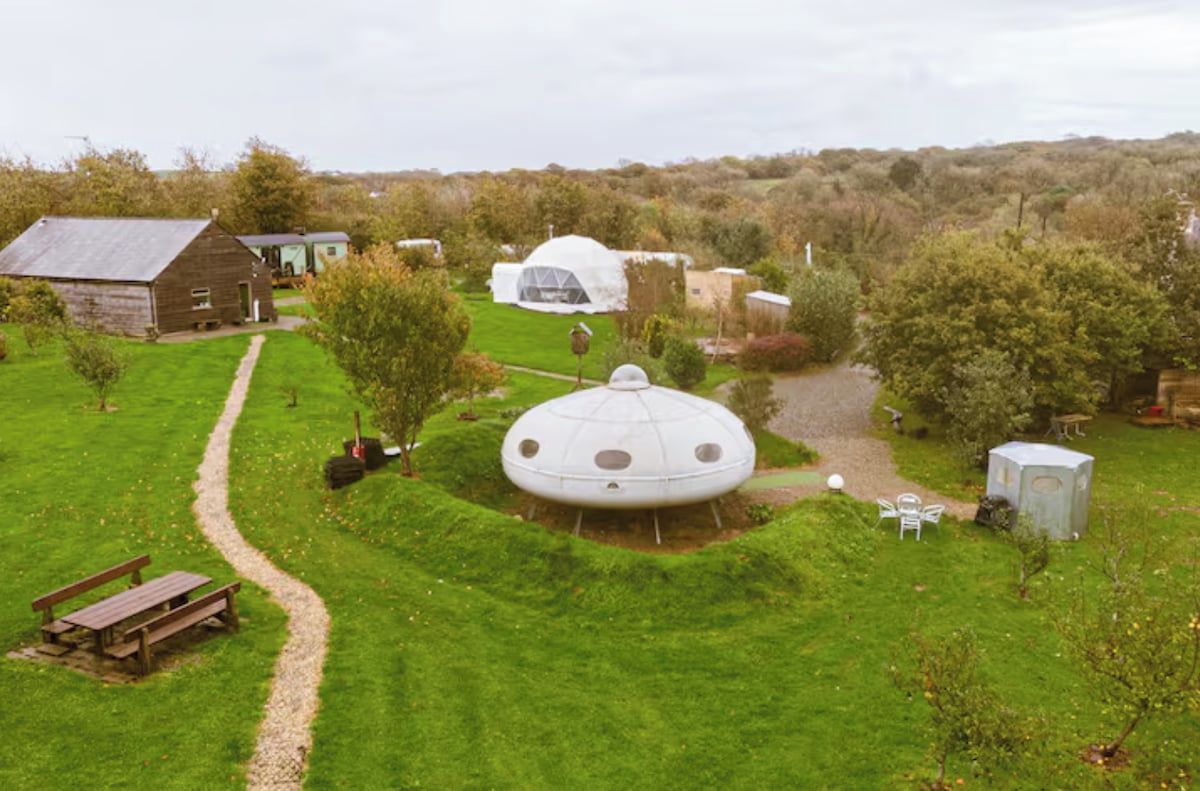 A rural setting with a large geodesic dome structure surrounded by a well-maintained lawn, a wooden barn, and a small shed.