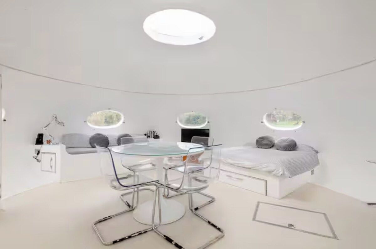 Un dormitorio moderno con una mesita de vidrio, una cama con cabecera blanca y dos lámparas fijadas a la pared.
