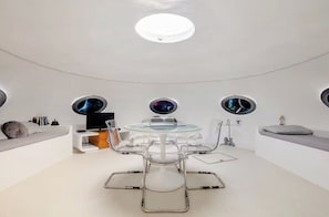 Una habitació moderna i circular amb una taula i cadires de vidre, sostre blanc i finestres rodones decoratives.