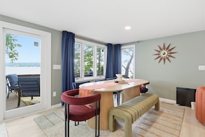 Un comedor con una mesa, sillas y un banco. Hay ventanas con cortinas, vistas al exterior y un reloj en la pared.