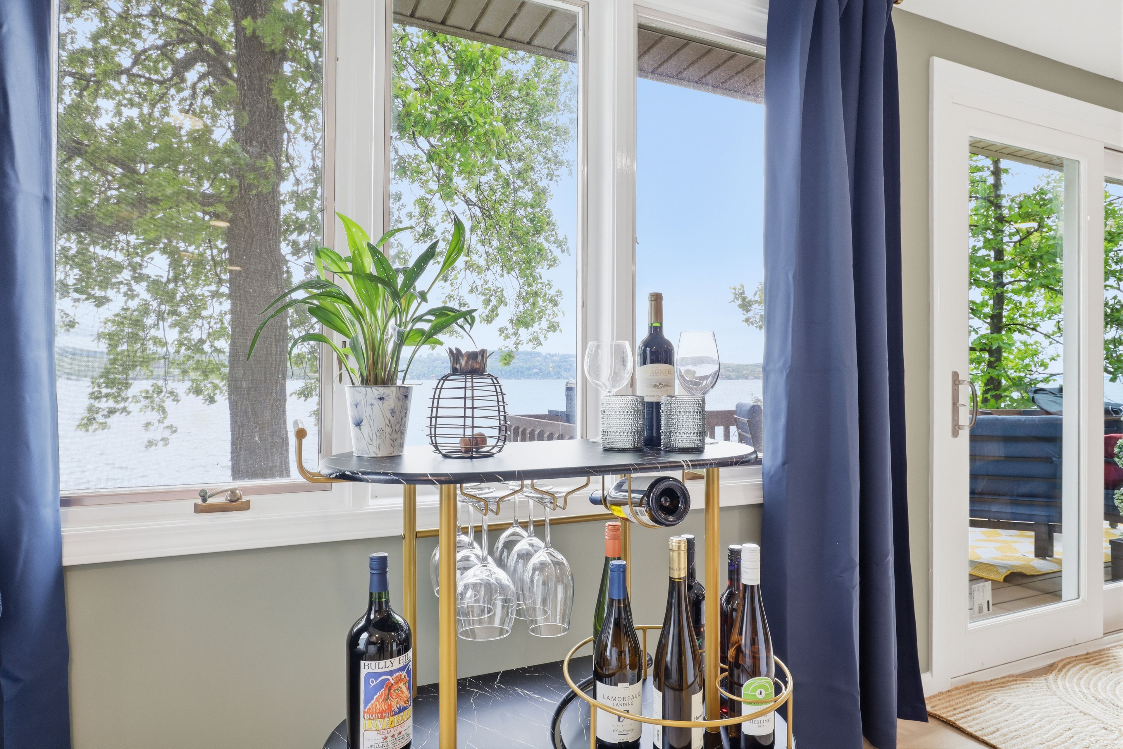 A table with wine bottles and glasses, a potted plant, and a view of trees through a window.