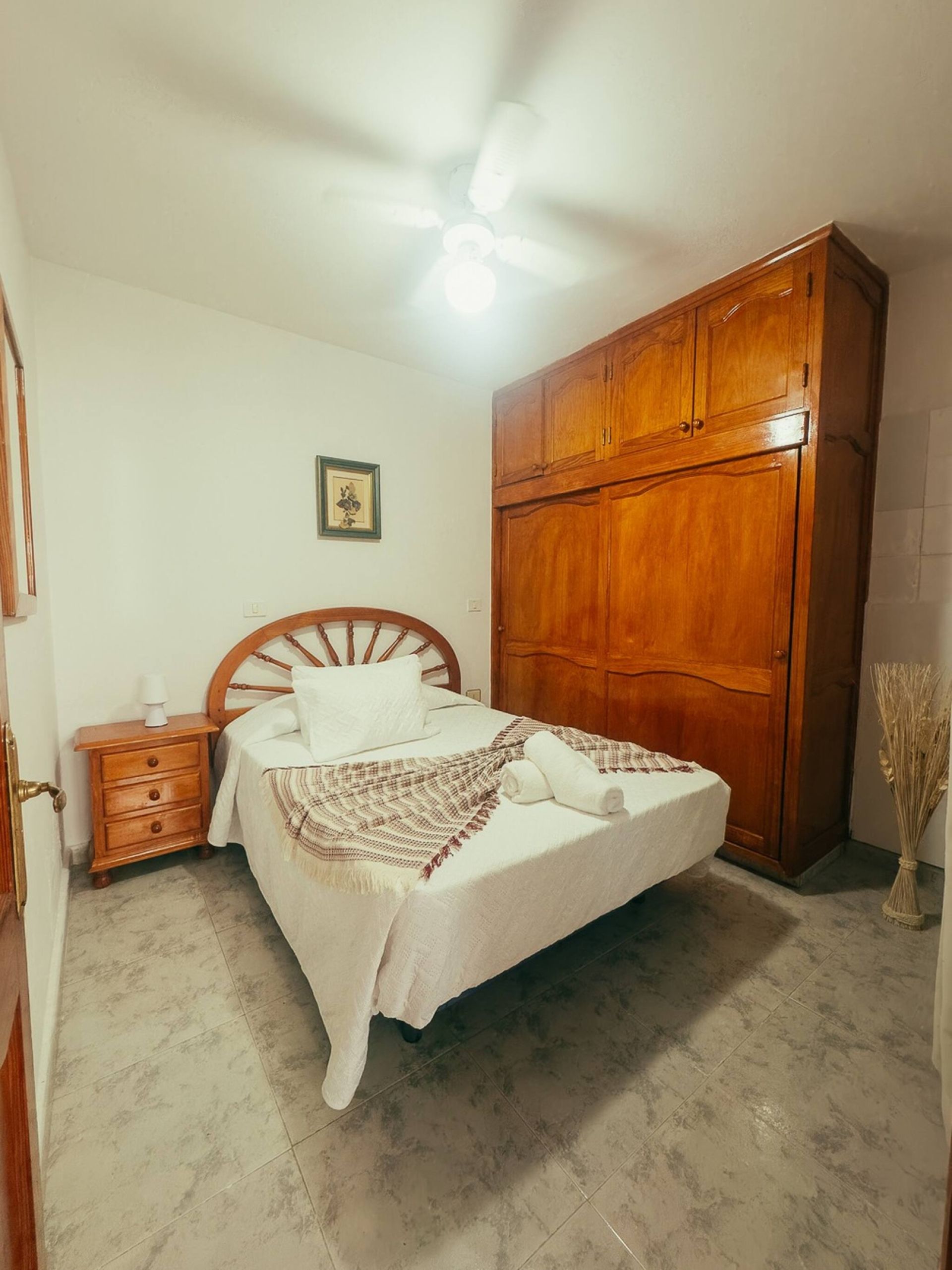 Une chambre à coucher comprenant un lit, une armoire en bois, une table de chevet et un ventilateur de plafond.