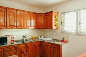 A kitchen with wooden cabinets, a microwave, a kettle, a toaster, and a window with blinds. - Apartment 2 | Aldea Blanca | 2 bedrooms | Hiking trails (Aldea Blanca)