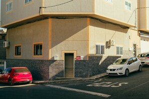 Ein Gebäude mit einer flachen Fassade, einer Tür mit einem ‚V‘-Schild und zwei parkenden Autos auf der Straße.