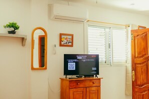 Een kamer met een televisie op een houten kast, een wandairconditioner, een raam met zonwering, een deur met een koperen handgreep, en een kleine potplant op een plank.