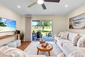A modern living room with a flat-screen TV, a sofa, a coffee table, and a view of a golf course through a sliding glass door. - Vista Verde East 105 (St. Petersburg)